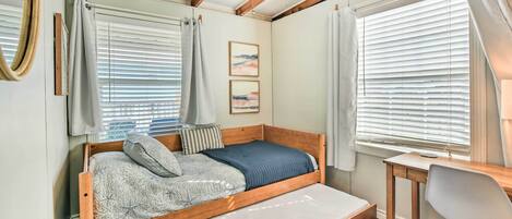 A bedroom with a wooden bed, a desk, and a chair. There are two windows with blinds and a ceiling fan.