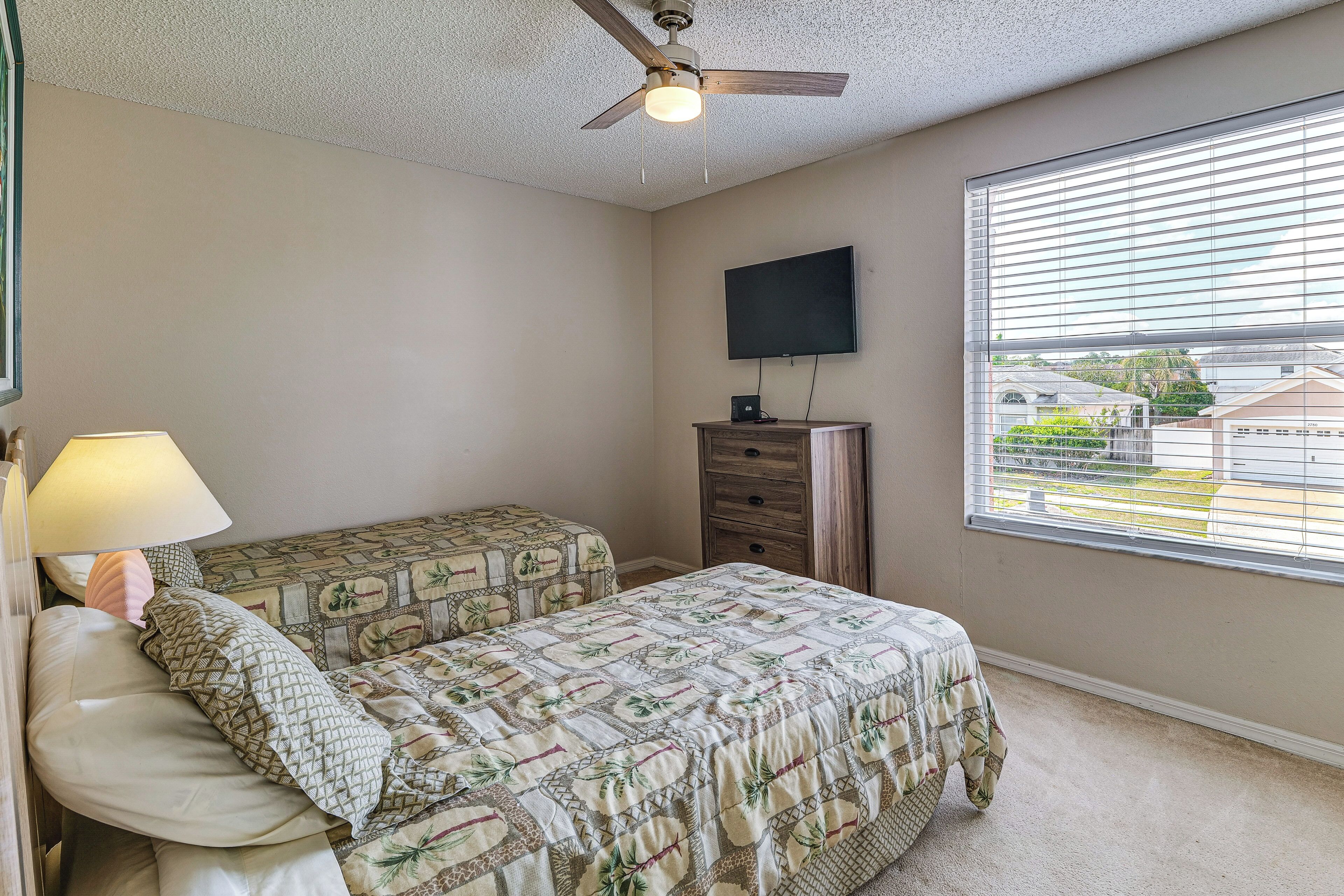 A bedroom with two beds, a ceiling fan, a TV, and a window with blinds.