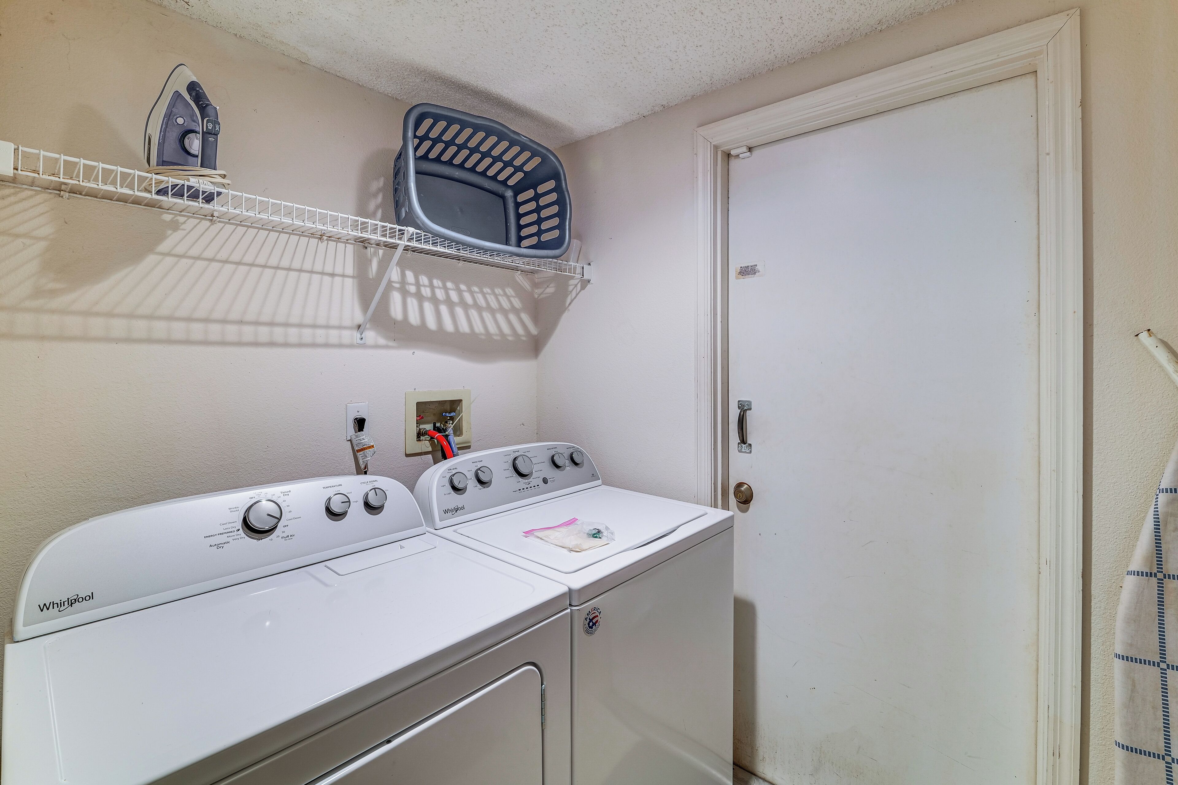 Un área de lavandería con lavadora y secadora Whirlpool, un estante con una cesta y una puerta.