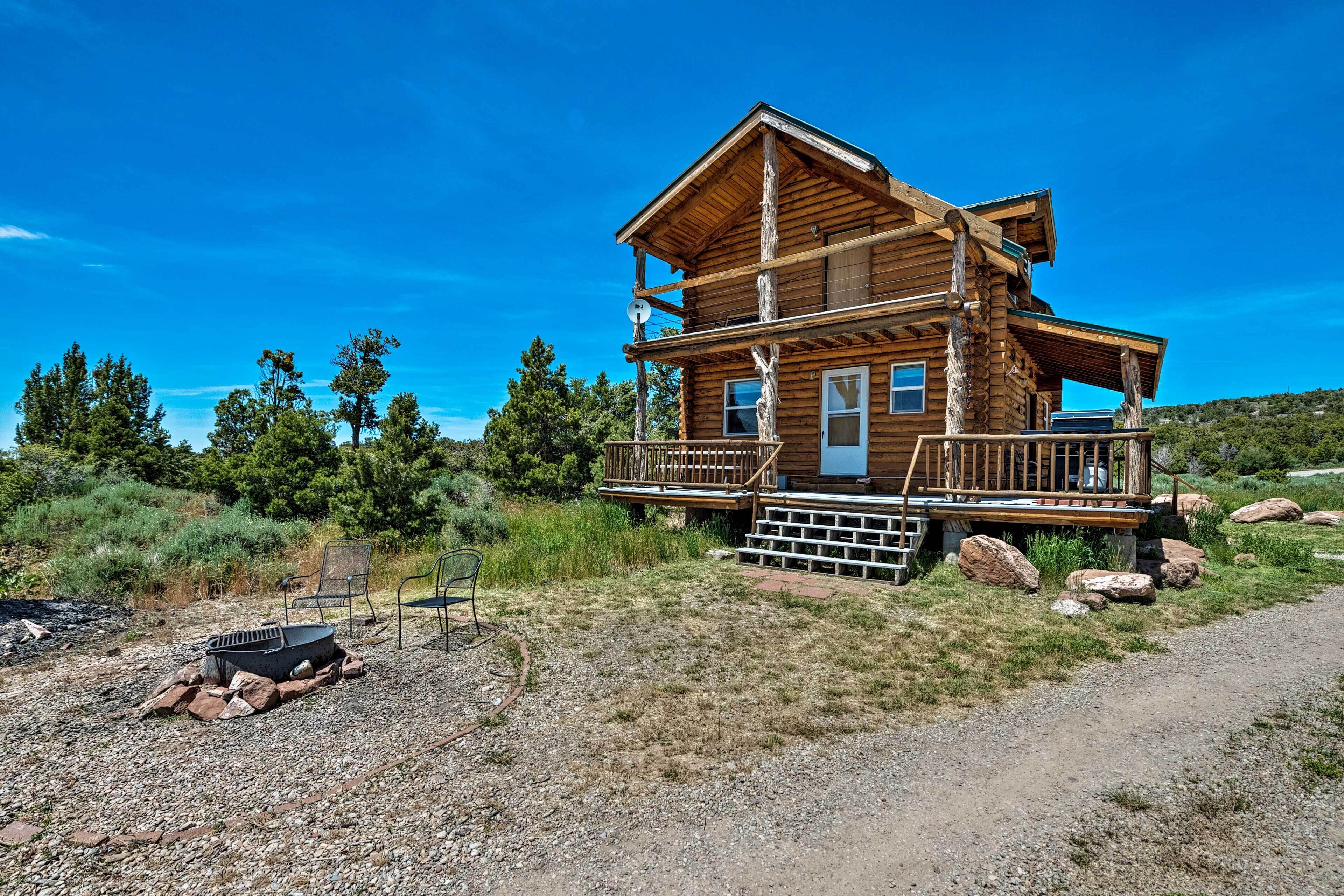 A two-story log cabin with a porch, surrounded by a gravel driveway and natural landscape.