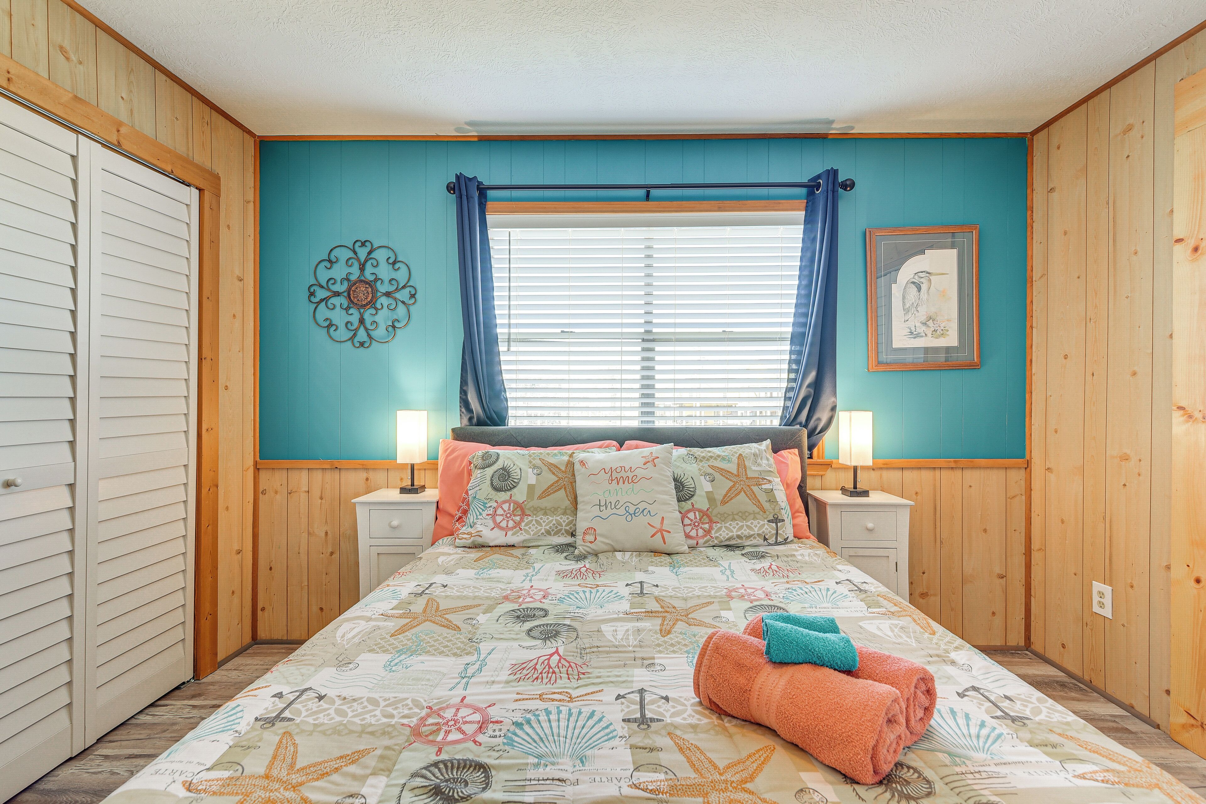 A bedroom with a bed, two bedside tables, a window with curtains, a wall decoration, and a framed picture.