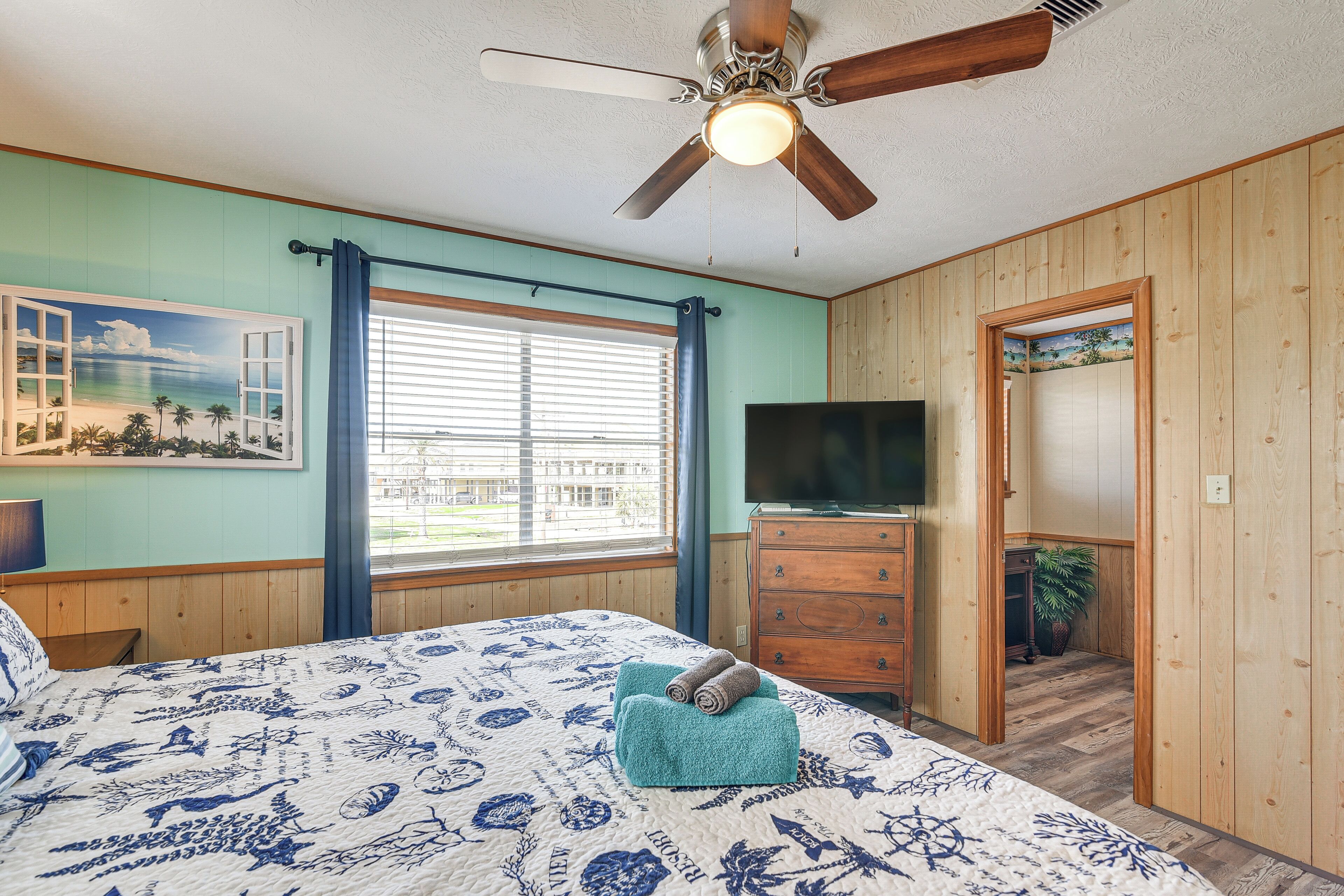 A bedroom with a bed, a ceiling fan, a window with blinds, a dresser, and a TV.