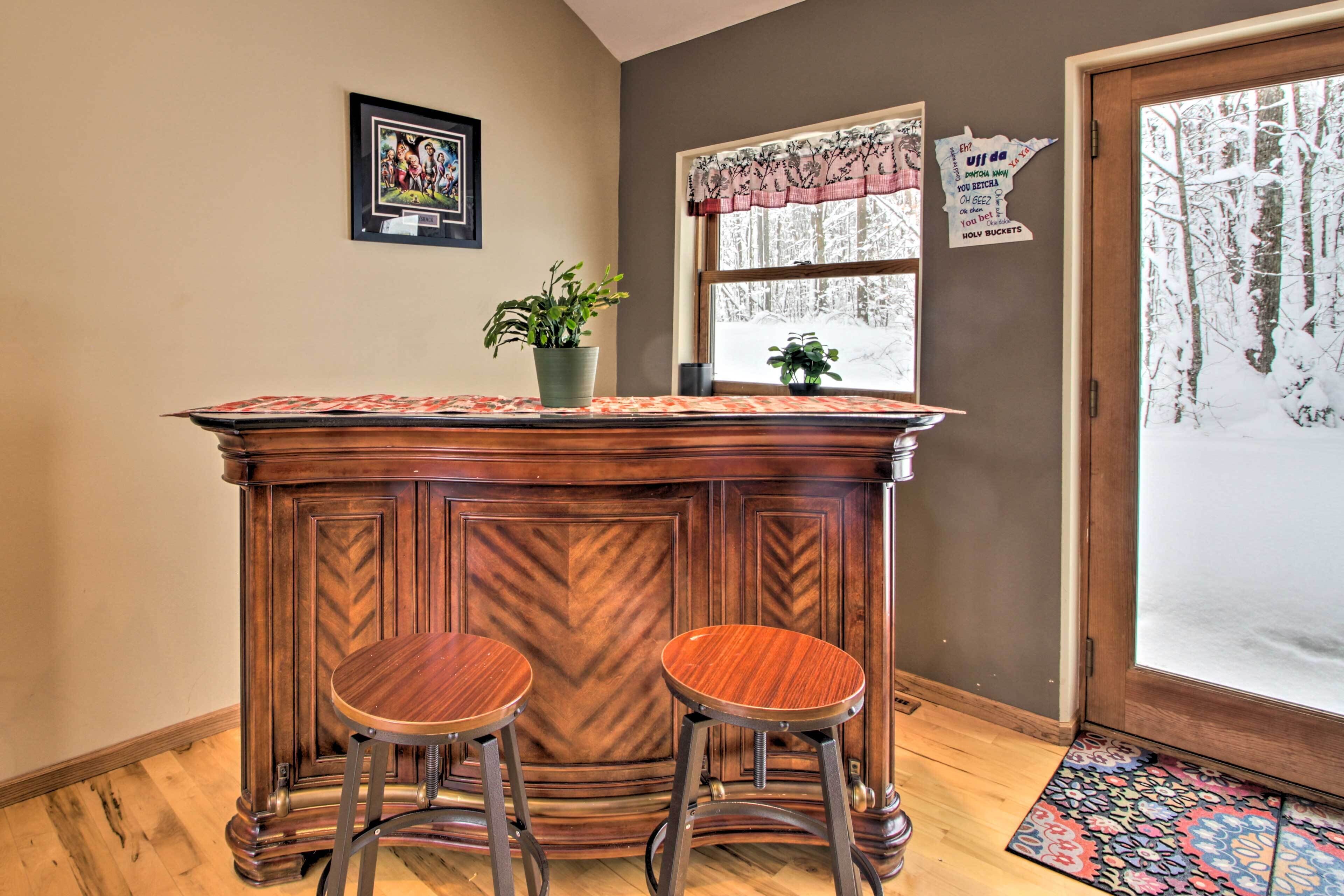 Un coin bar avec deux tabourets, une plante en pot, un tableau encadré, une fenêtre donnant sur des arbres et une porte avec un panneau en verre dépoli.