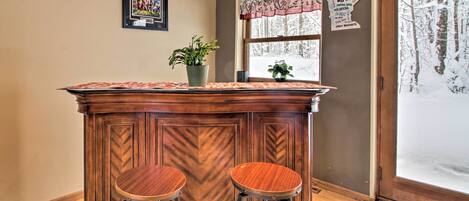 Un coin bar avec deux tabourets, une plante en pot, un tableau encadré, une fenêtre donnant sur des arbres et une porte avec un panneau en verre dépoli.