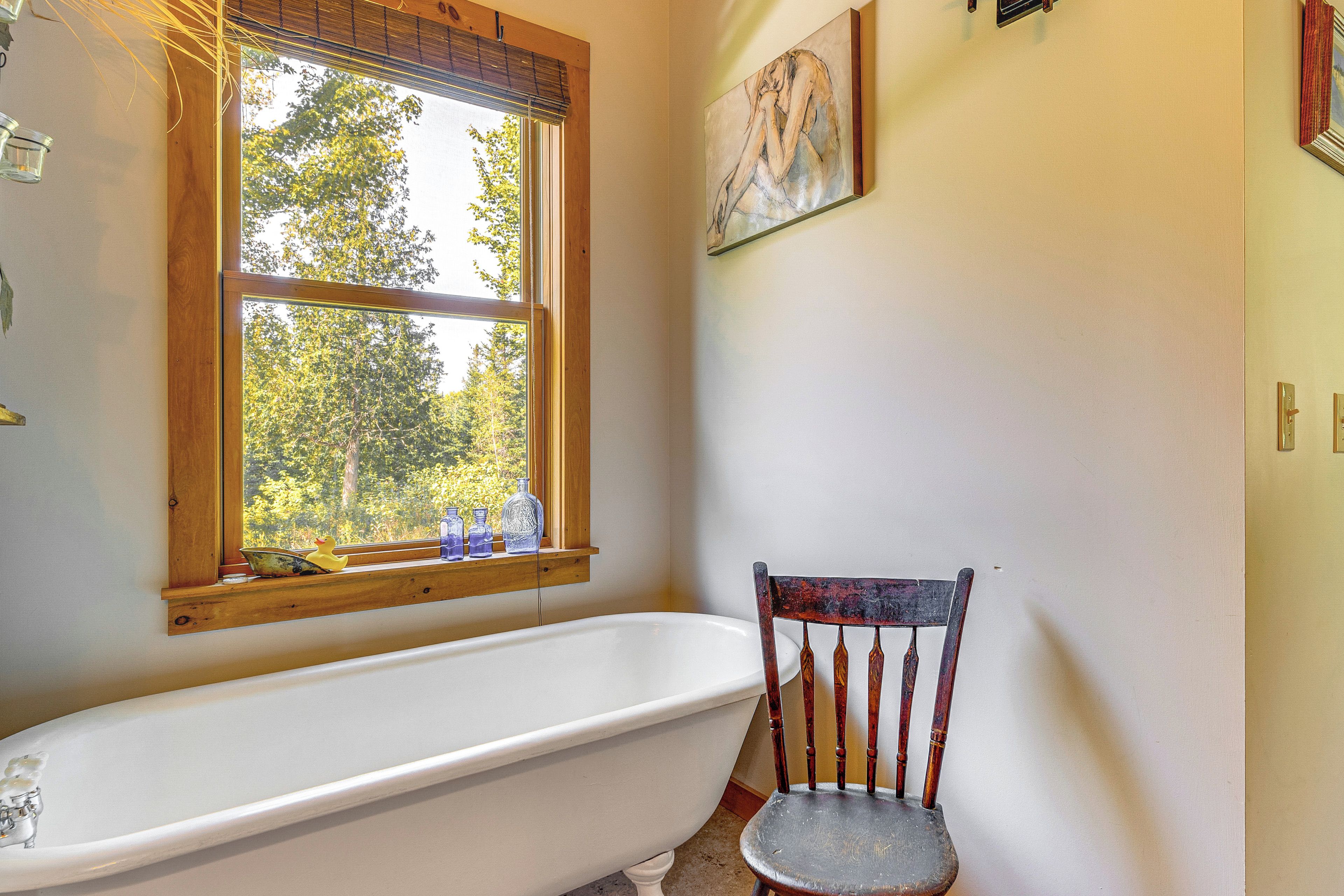 A bathroom with a white bathtub, a wooden chair, a window with a view of trees, and a painting on the wall.