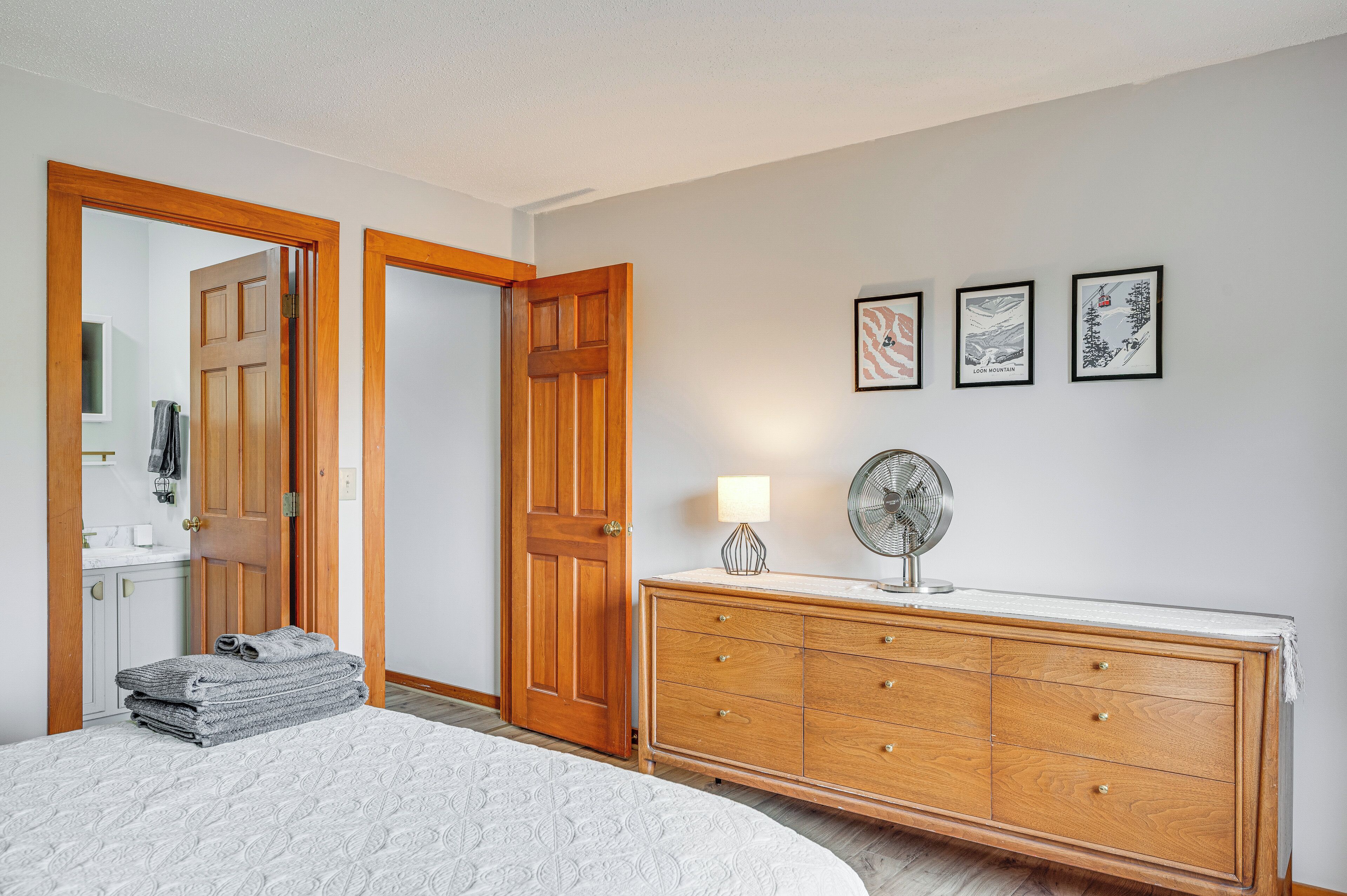 A bedroom with a bed, a wooden dresser, a fan, and three framed pictures on the wall.