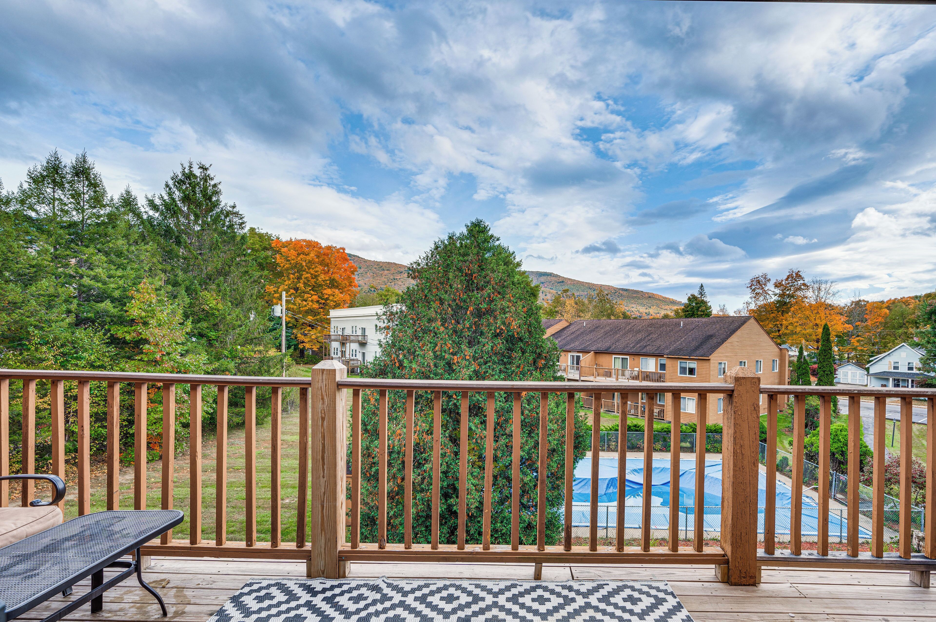 A wooden deck with a metal table and bench, overlooking a swimming pool and houses in a hilly area.