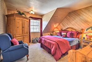 A cozy bedroom with a sloped ceiling, a wooden wardrobe, a bed with a red bedspread, a blue armchair, and a lamp on the nightstand.