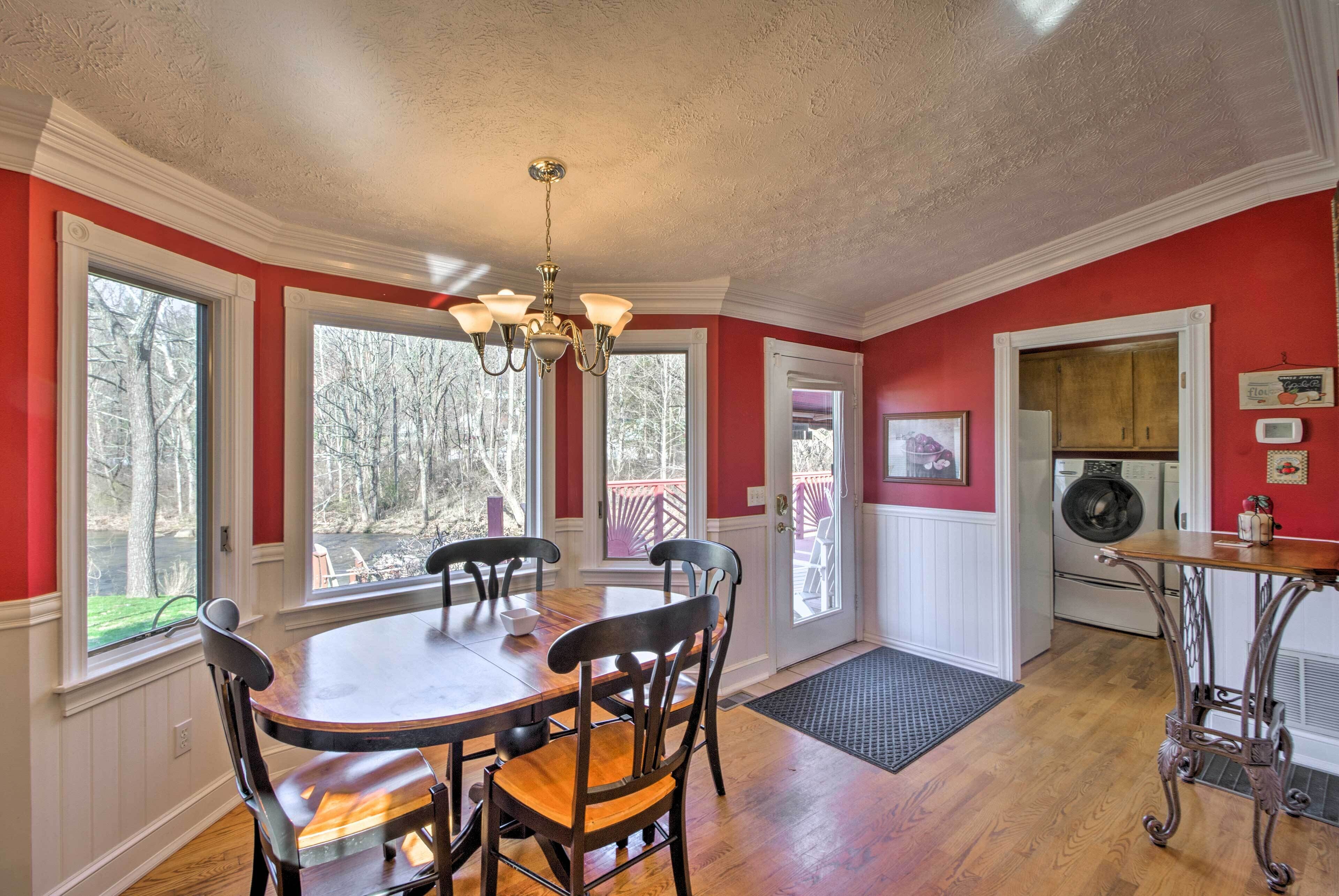 A dining area with a wooden table and chairs, a chandelier, and large windows with a view of trees.