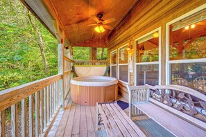 Een houten terras met een jacuzzi, een plafondventilator en uitzicht op het omliggende bos.