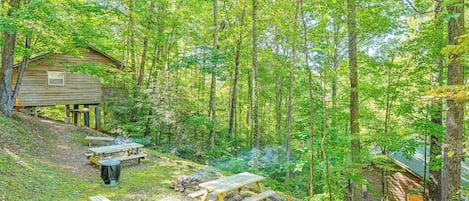 Un chalet rustique doté d’une terrasse en bois et de tables de pique-nique, situé dans une zone boisée.