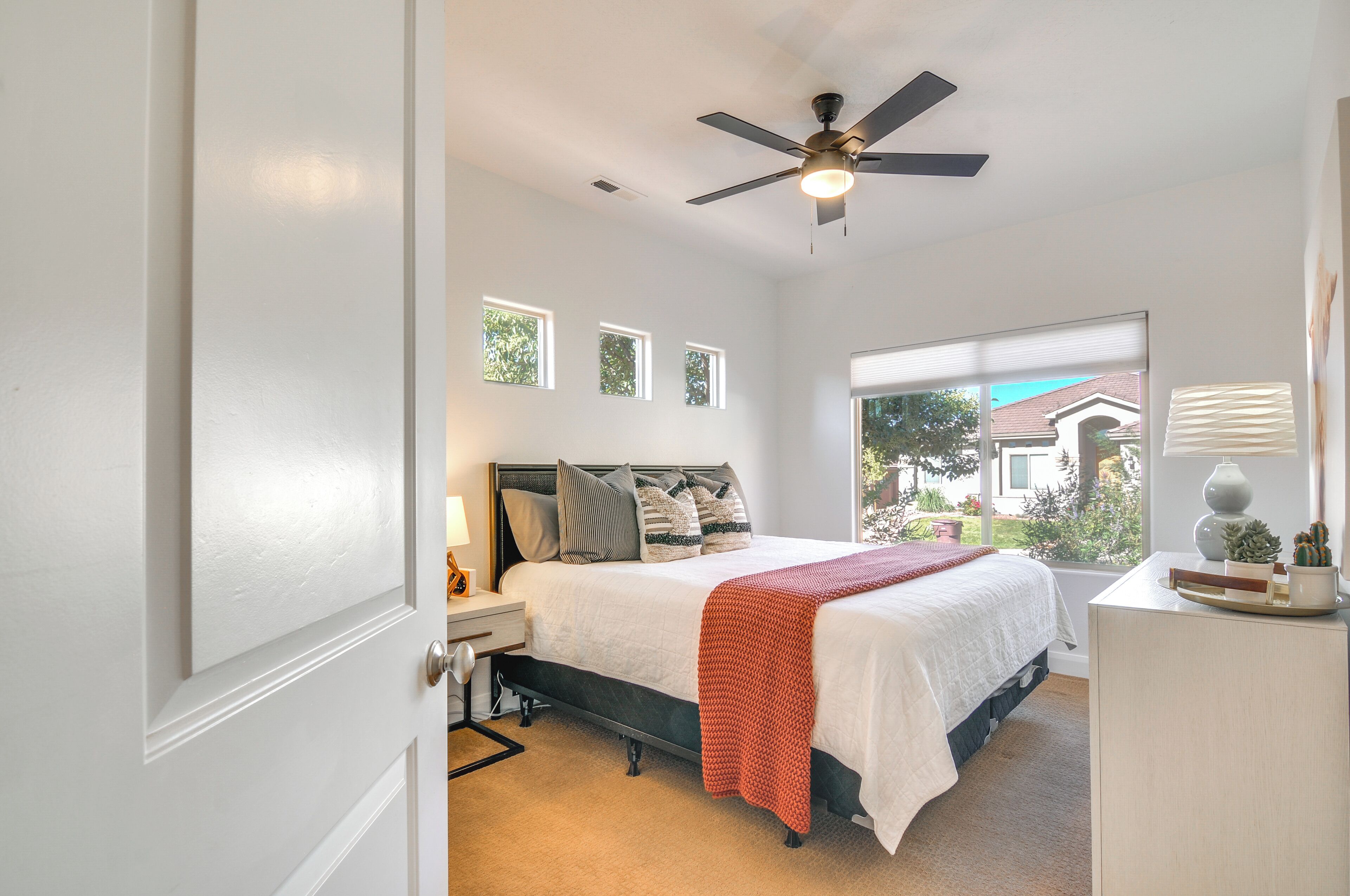 A bedroom with a bed, bedside table, lamp, ceiling fan, window, and view of the outside.