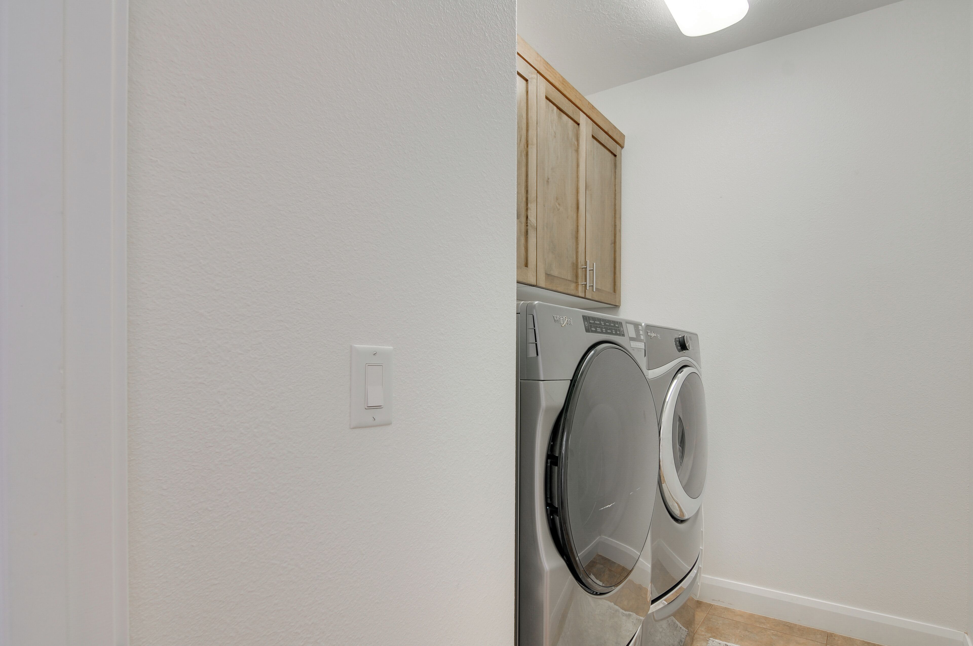 A laundry room with a front-loading washing machine, wooden cabinets above, and a white wall.