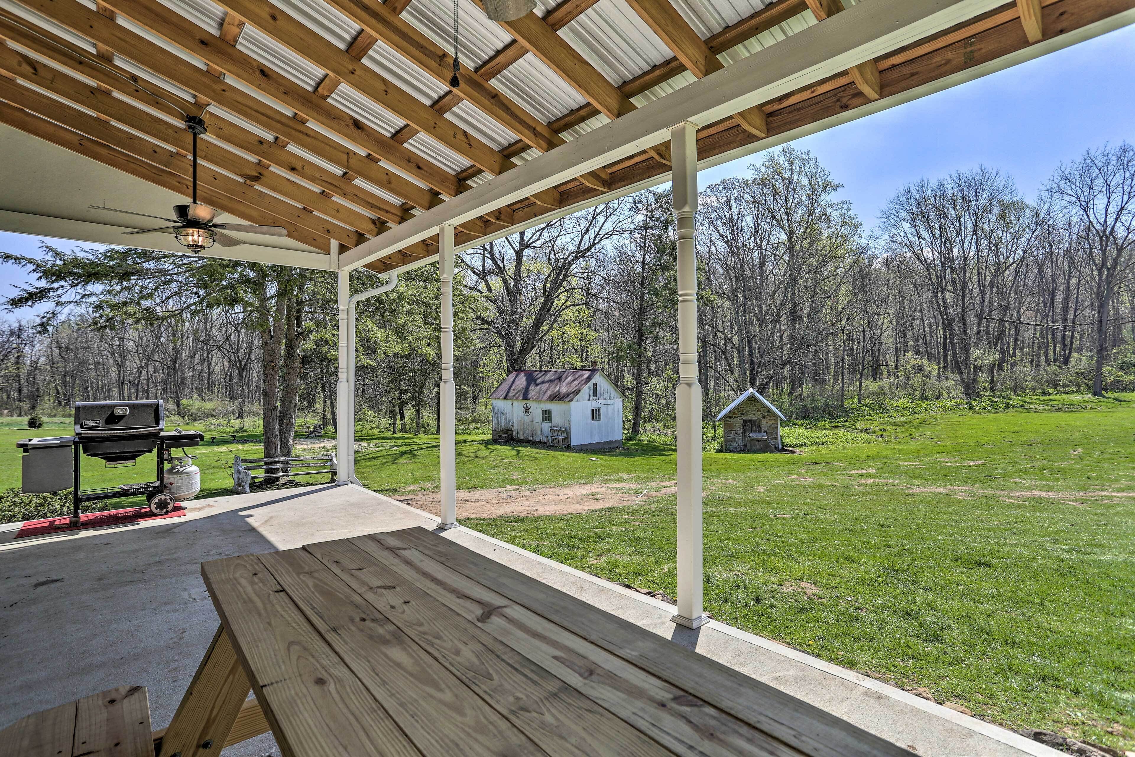 A covered patio with a ceiling fan, a grill, and a view of a small white house in a wooded area.