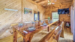 A wooden cabin interior with a long dining table set for four, a television mounted on the wall, and a ceiling fan. - Spacious Cabin on Dale Hollow Lake w/ Hot Tub! (Albany)