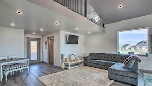 Ein geräumiges Wohnzimmer mit grauem Sofa, einem Esstisch mit Stühlen, einem großen Fenster mit Blick und Einbauleuchten.