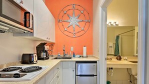 A compact kitchen with a stove, toaster, and coffee maker. A stainless steel refrigerator is next to the sink, which has a soap dispenser and a paper towel holder. A large compass decoration adorns the orange wall. - Everglades Studio W/marina View, Patio+pool Access (Naples)