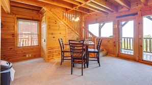 A wooden cabin interior with a dining table, chairs, a staircase, and large windows.
