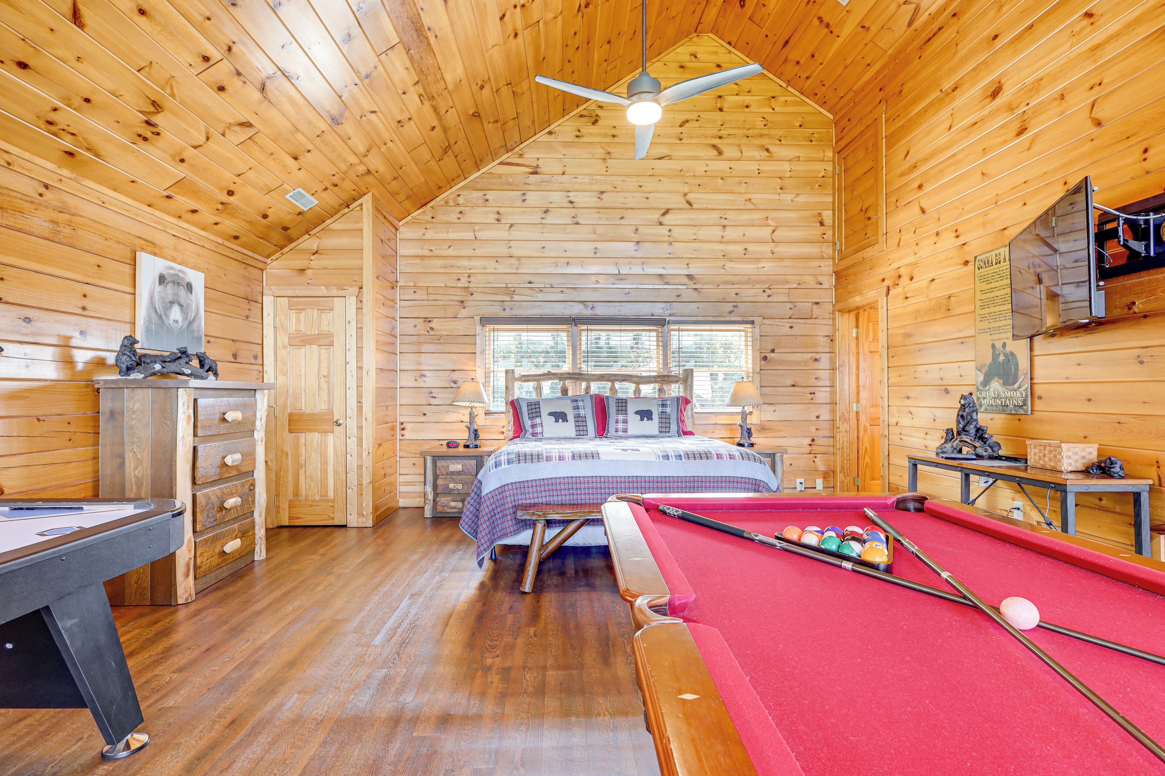 A wooden cabin interior with a pool table, a billiards table, a bed with a checkered bedspread, and a ceiling fan.