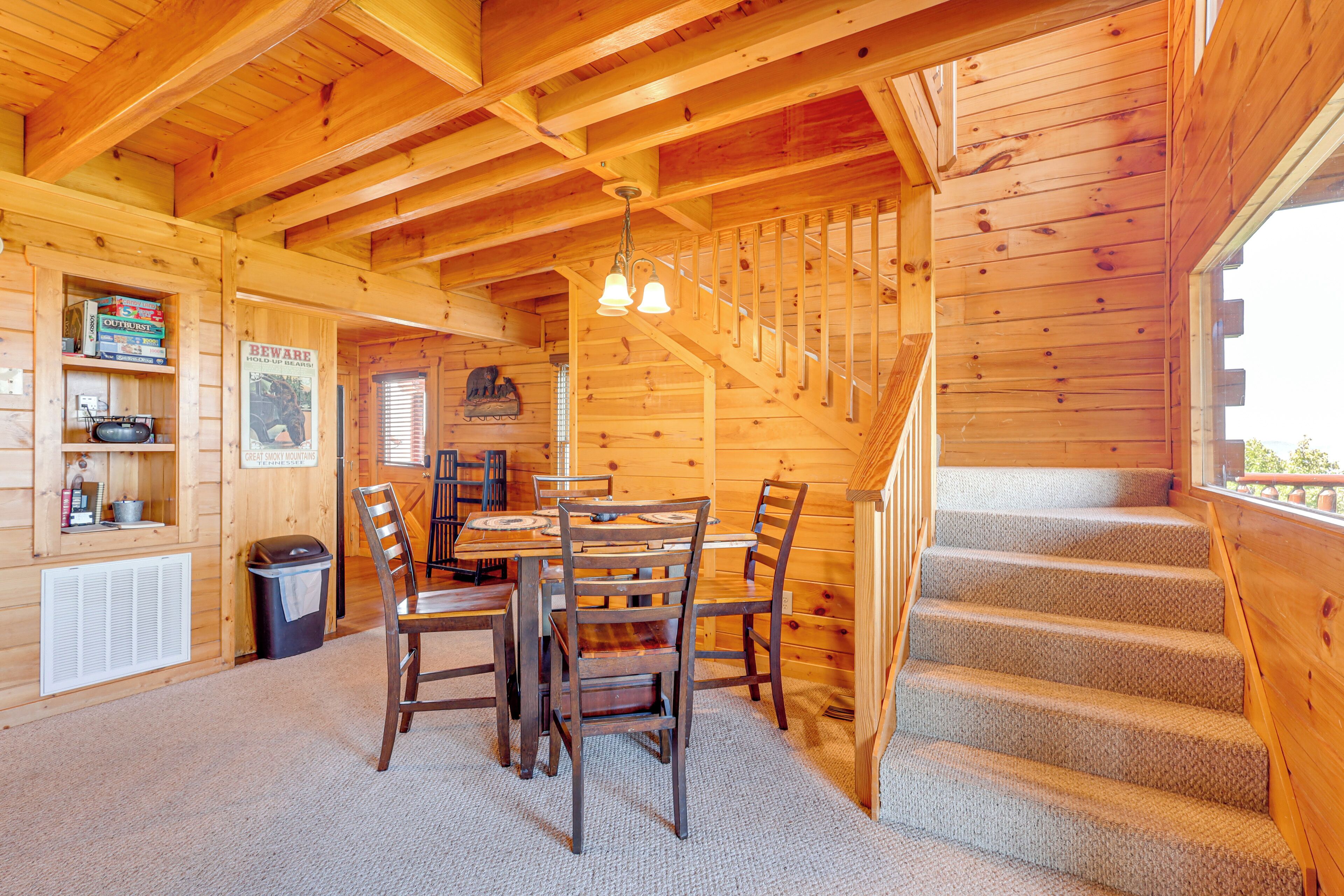 A wooden cabin interior with a dining area, a staircase, and a bookshelf with books and a radio.