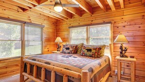 A wooden cabin bedroom with a bed, two windows with blinds, a ceiling fan, and a nightstand with a lamp.