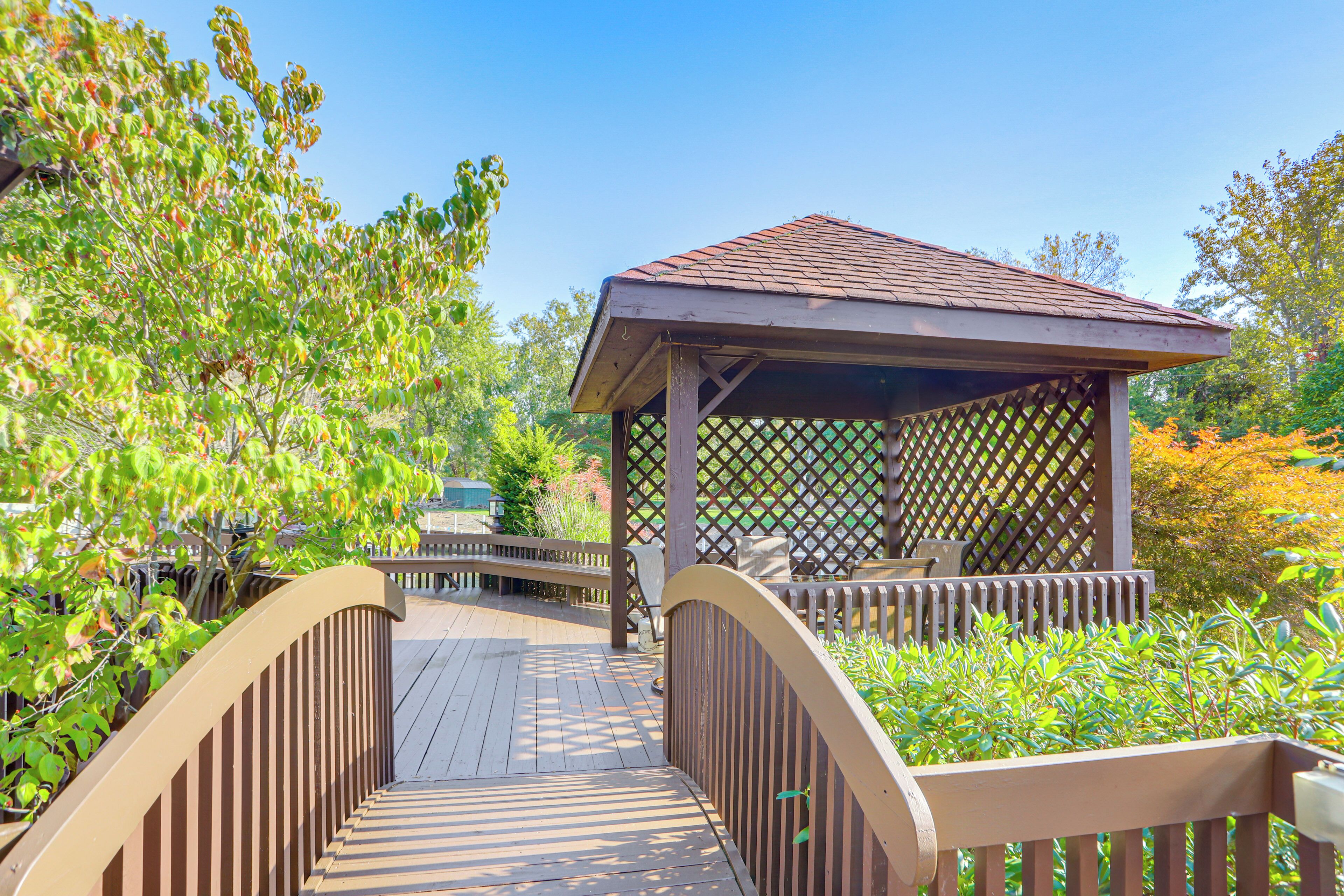 A wooden bridge leading to a gazebo with a lattice roof, surrounded by lush greenery.