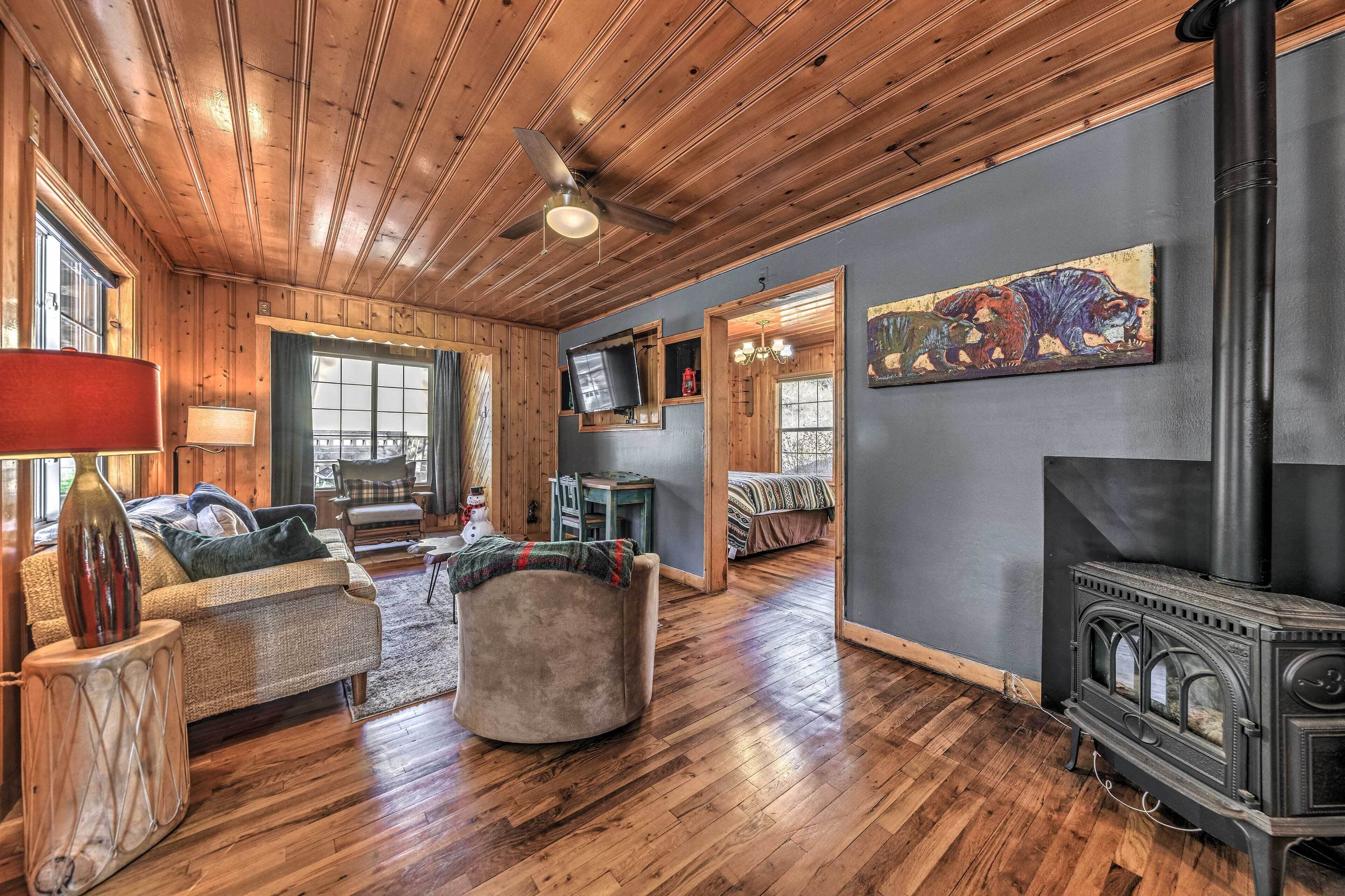 L’intérieur chaleureux d’un chalet avec un plafond en bois, une cheminée, un téléviseur fixé au mur et un coin salon confortable avec un canapé et un pouf.