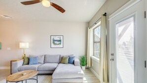 Ein modernes Wohnzimmer mit einem grauen Ecksofa, einem Holztisch, einem Deckenventilator, einem gerahmten Landschaftsbild und einem großen Fenster mit Jalousien.