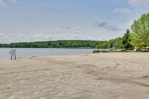 Uma praia arenosa com uma placa, um lago e uma colina arborizada ao fundo.