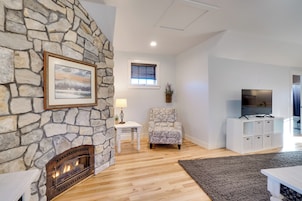A living room with a stone fireplace, a framed picture, a patterned armchair, a white side table, a TV stand, and a gray rug.