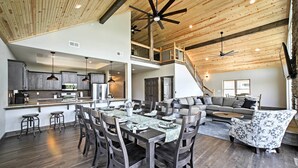 A spacious living area with a high ceiling, wooden beams, and a dining table set for six. - Lavish Retreat w/ Decks, Steps to Table Rock Lake! (Lampe)
