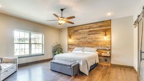 A bedroom with a wooden accent wall, a ceiling fan, a large window, a bed, a bench, and a nightstand with a lamp. - Lavish Retreat w/ Decks, Steps to Table Rock Lake! (Lampe)