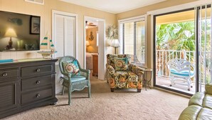 A living room with a dark wooden dresser, two patterned chairs, a television, and a sliding glass door leading to a balcony with plants.