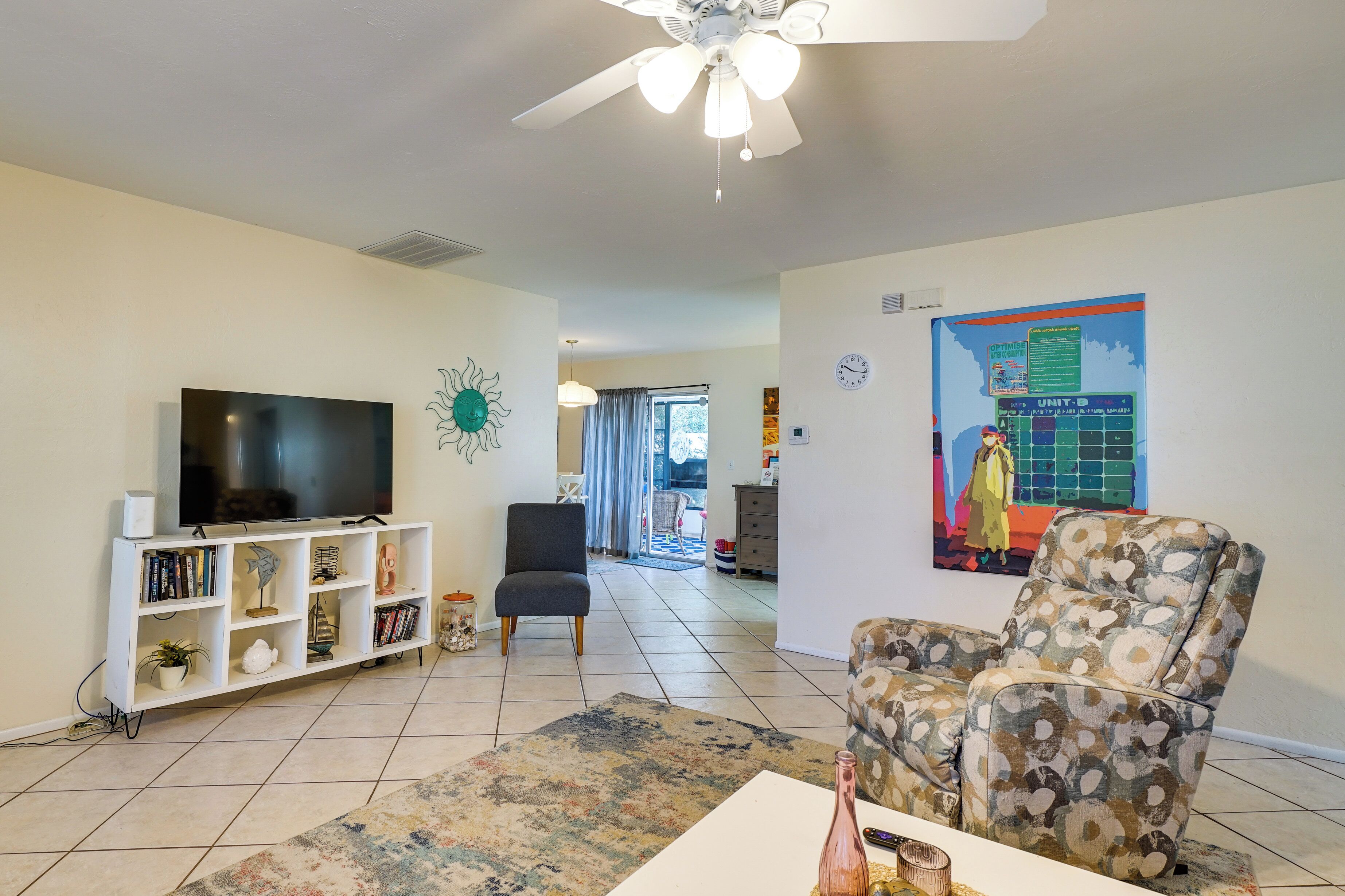 A living room with a flat-screen TV, a bookshelf, a patterned armchair, and a ceiling fan.