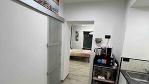 A narrow hallway with a kitchen area on the right, a bed in the background, and a door on the left. - CaBi's Dreams (Napoli)
