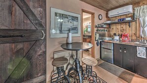 Cozinha com porta de madeira, um quadro emoldurado, uma mesa redonda pequena com banquetas e uma geladeira.
