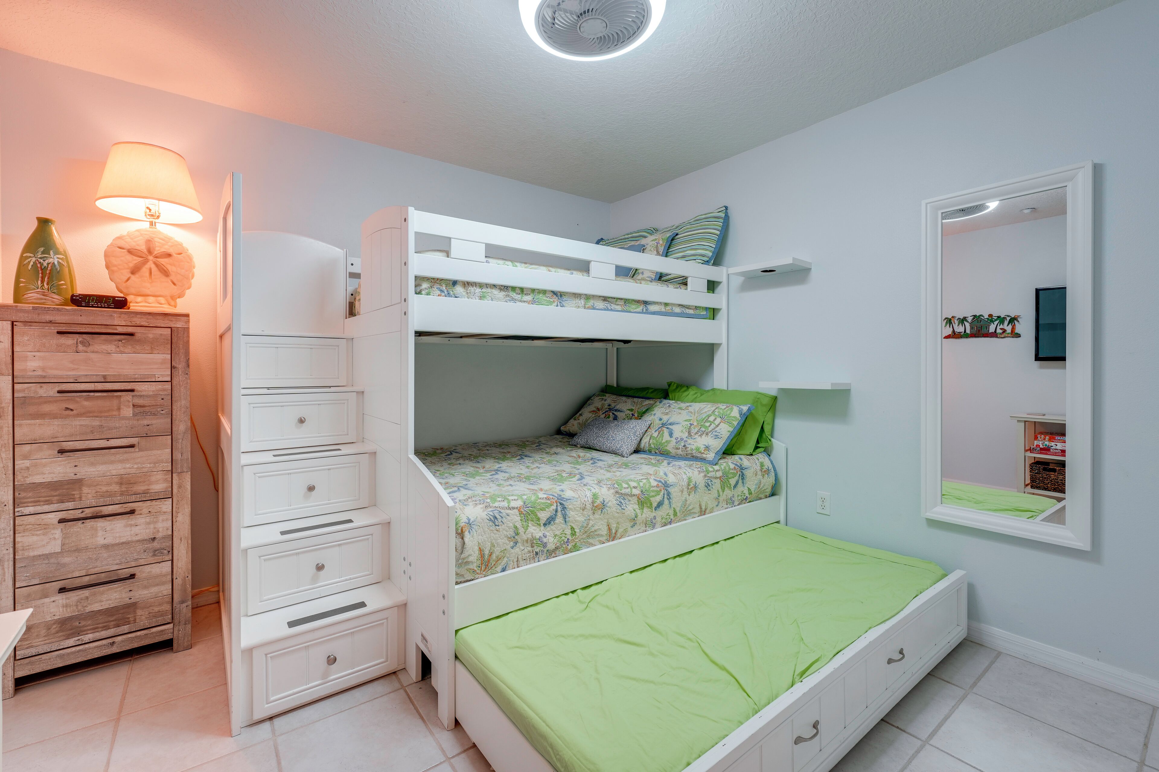 Ein Etagenbett mit einer grünen Matratze, eine Holzkommode, eine Lampe und ein Spiegel an der Wand.