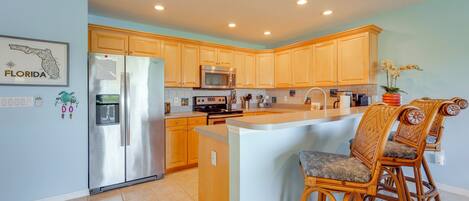 A kitchen with wooden cabinets, a stainless steel refrigerator, a microwave, and a bar area with wicker chairs.
