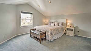 A bedroom with a sloped ceiling, a large bed with patterned bedding, a nightstand with a lamp, and a window with a view of buildings. - Laramie Home w/ Backyard < 1 Mi to Univ of Wyoming (Laramie)