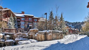 A snow-covered outdoor area with a wooden deck, a large rock formation, and a building with multiple balconies and windows. - Solitude Mountain Slopeside Ski Condo w/ Hot Tub! (Solitude)