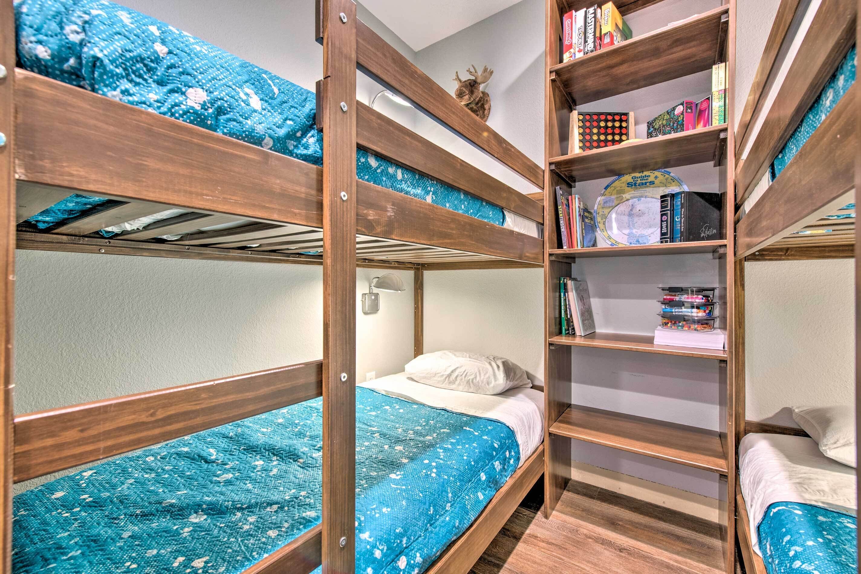 A bunk bed room with a blue patterned bedspread, a bookshelf with various books and a decorative item on the wall, and a small table with a lamp.