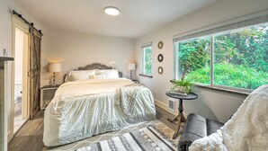 A bedroom with a large bed, bedside tables, a window with a view of greenery, and a door leading to another room. - Farmhouse w/ Private Hot Tub < 1 Mile to Birch Bay (Blaine)