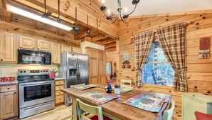 A wooden cabin interior with a kitchen featuring stainless steel appliances, a dining table with placemats, and a window with plaid curtains. - Waynesville Cabin w/ Covered Deck & Fire Pit! (Waynesville)