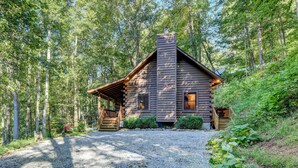 A rustic log cabin with a chimney, a wooden porch, and a gravel driveway surrounded by dense forest. - Waynesville Cabin w/ Covered Deck & Fire Pit! (Waynesville)
