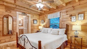 A wooden cabin bedroom with a bed, a ceiling fan, a window with blinds, a mirror, and a small table with a lamp. - Waynesville Cabin w/ Covered Deck & Fire Pit! (Waynesville)