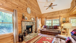 A cozy cabin interior with a stone fireplace, leather sofa, and a large window offering a forest view. - Waynesville Cabin w/ Covered Deck & Fire Pit! (Waynesville)