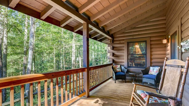 A wooden deck with a railing, a wicker chair, a table, and a hanging light fixture. - Waynesville Cabin w/ Covered Deck & Fire Pit! (Waynesville)