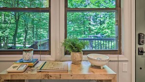 A wooden table with a bowl, a potted plant, and books by a window with a view of trees and a deck.