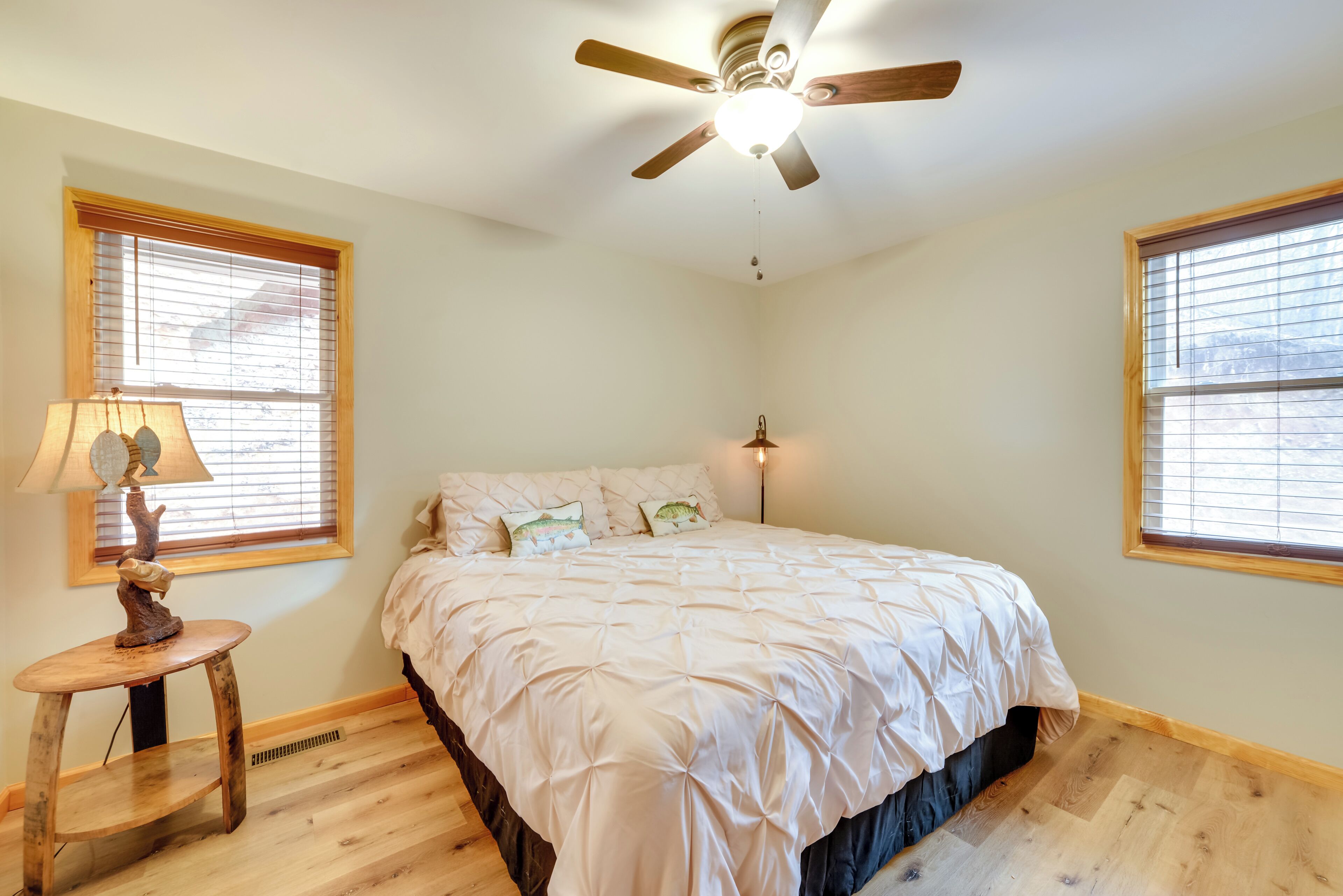 A bedroom with a bed, two windows, a ceiling fan, and a small table with a lamp.