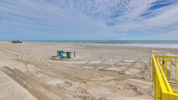 Una playa con una baranda amarilla, una torre de salvavidas azul y marcas de neumáticos en la arena.
